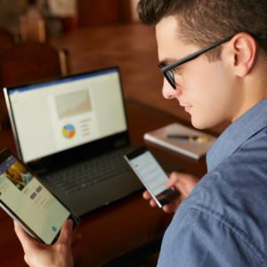 Attractive man in glasses working with multiple electronic internet devices. Freelancer businessman has laptop and smartphone in hands and laptop on table with charts on screen. Multitasking theme